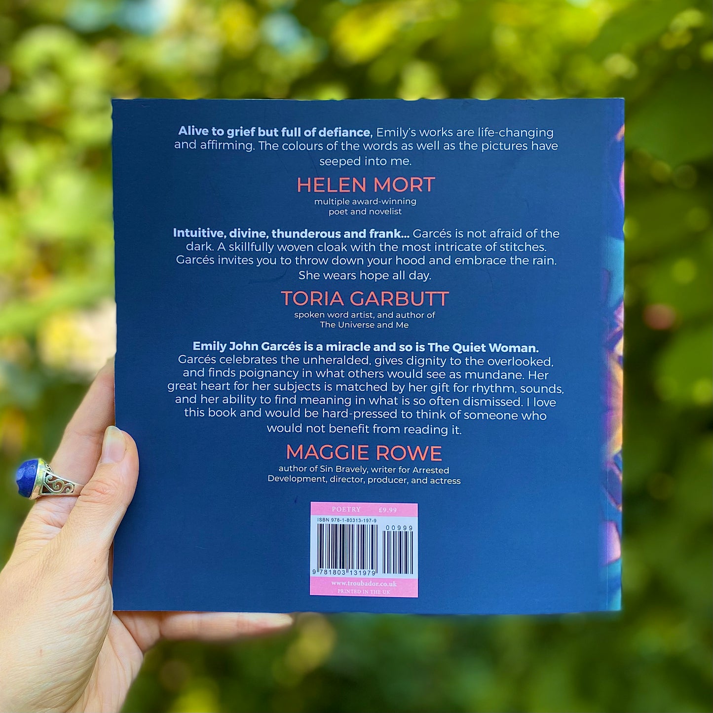 woman's hand holding The Quiet Woman book to show the back cover in dark blue with testimonials from authors, poets, and producers