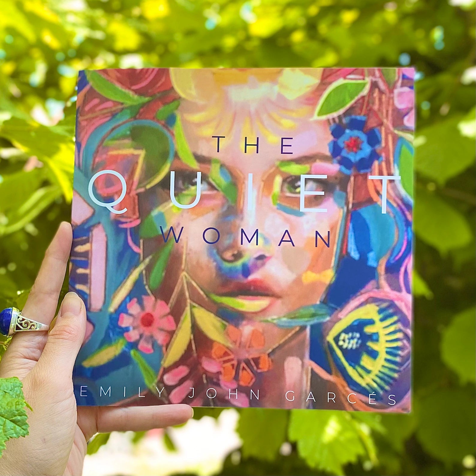 woman's hand holding The Quiet Woman book showing its colourful folk-artsy front cover, with verdant green leaves of a bush in the background.